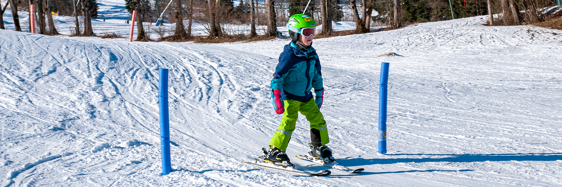 Skikurs Kind fährt bei schönem Wetter mit den Ski einen kleinen Hügel hinunter durch ein aufgestelltes Tor